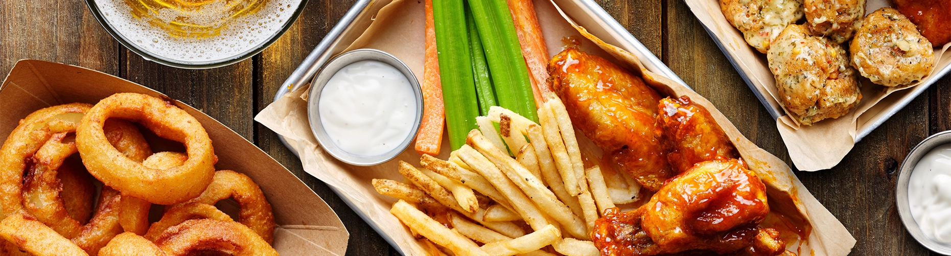 A top-down view of a variety of bar foods on a wooden table, including a tray of buffalo wings, French fries, and celery sticks, a container of onion rings, and a glass of beer.