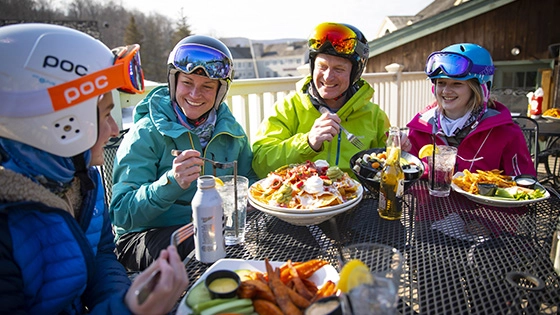 A smiling family of four in ski gear sits at an outdoor table, sharing a large plate of nachos and other food after a day on the slopes.