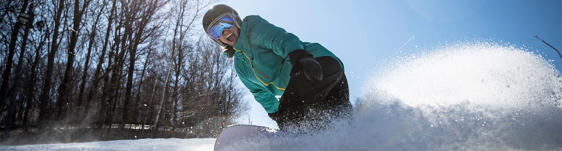 A smiling snowboarder, wearing a helmet and goggles, carves down a snowy slope, kicking up a large spray of powder with the bright sun behind them.