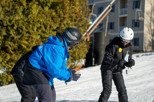 Two ski instructors are on a snowy slope, with one in a blue jacket demonstrating a technique to another instructor in a black jacket. A ski lift and a building are visible in the background.