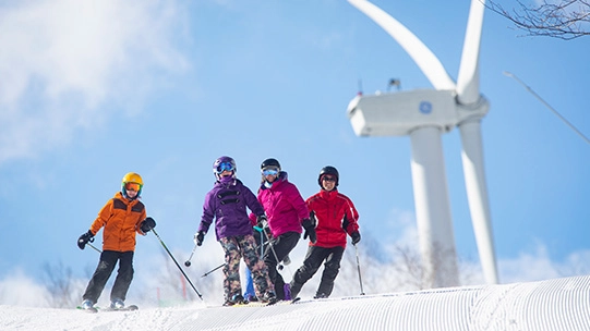 Four young skiers in colorful winter gear stand on a groomed snowy slope, smiling towards the camera. A large white wind turbine is prominently featured on the hill behind them.