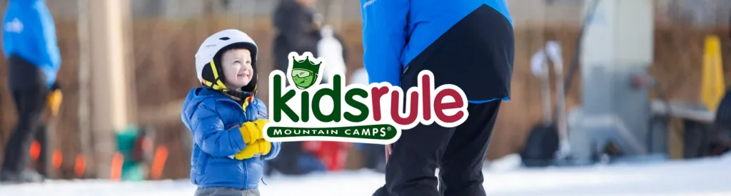 A happy young child in a blue jacket and white helmet looks up at a ski instructor standing in front of them. The KidsRule Mountain Camps logo is overlaid on the image.
