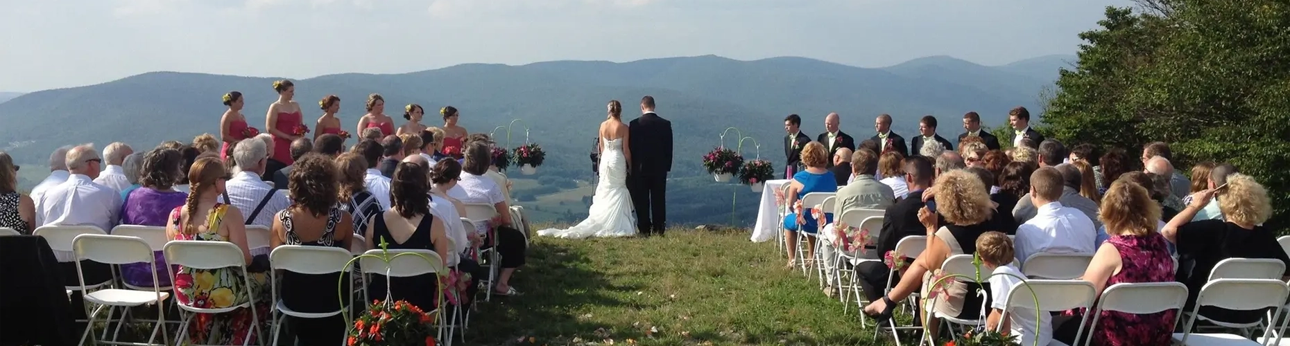 An outdoor wedding ceremony on a mountaintop, with the bride and groom standing on a grassy hill with their backs to a scenic mountain range. Guests are seated in rows of white chairs on either side.