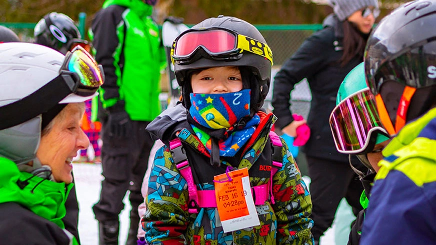 A close-up of a smiling toddler in ski gear, wearing a helmet, goggles, and a colorful face covering, standing among other skiers and instructors.