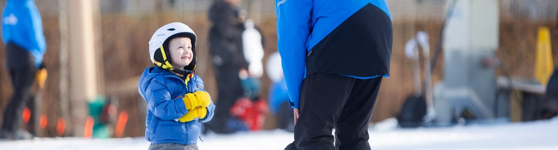 A happy young child in a blue jacket and white helmet looks up at a ski instructor standing in front of them. The KidsRule Mountain Camps logo is overlaid on the image.