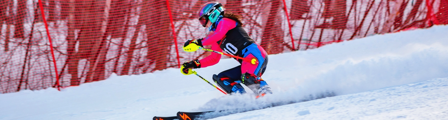 A skier in a brightly colored race suit and helmet is carving aggressively down a snowy racecourse, kicking up powder next to red safety netting. The image is a banner for the Jiminy Peak Race Teams.