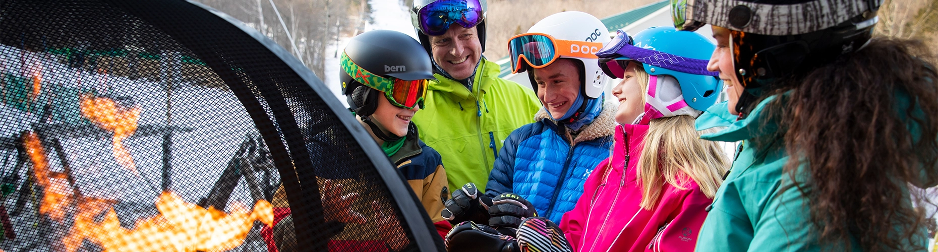 A group of smiling skiers and snowboarders in helmets and colorful gear gathers around a warm fire pit, set as the website banner for Snowsports Levels.