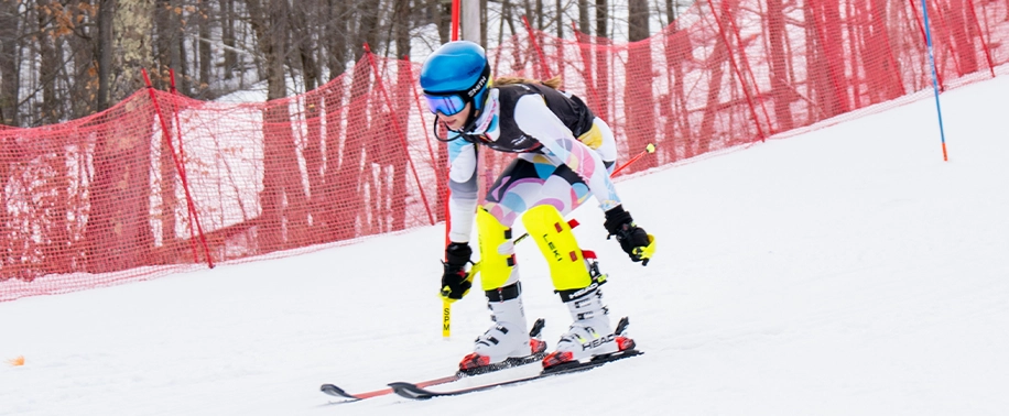 A young skier in a colorful race suit and bright yellow leg pads is speeding down a snowy slope, making a turn past red safety fencing for the U8/U10 Interclub Program.
