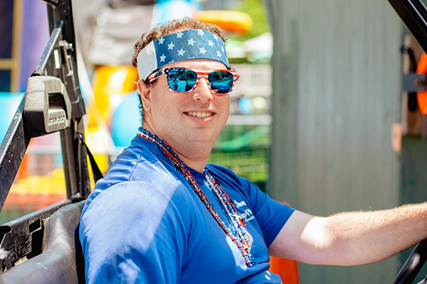 Smiling Jiminy Peak summer employee wearing American flag sunglasses, star bandana, and beaded necklace seated in a resort vehicle
