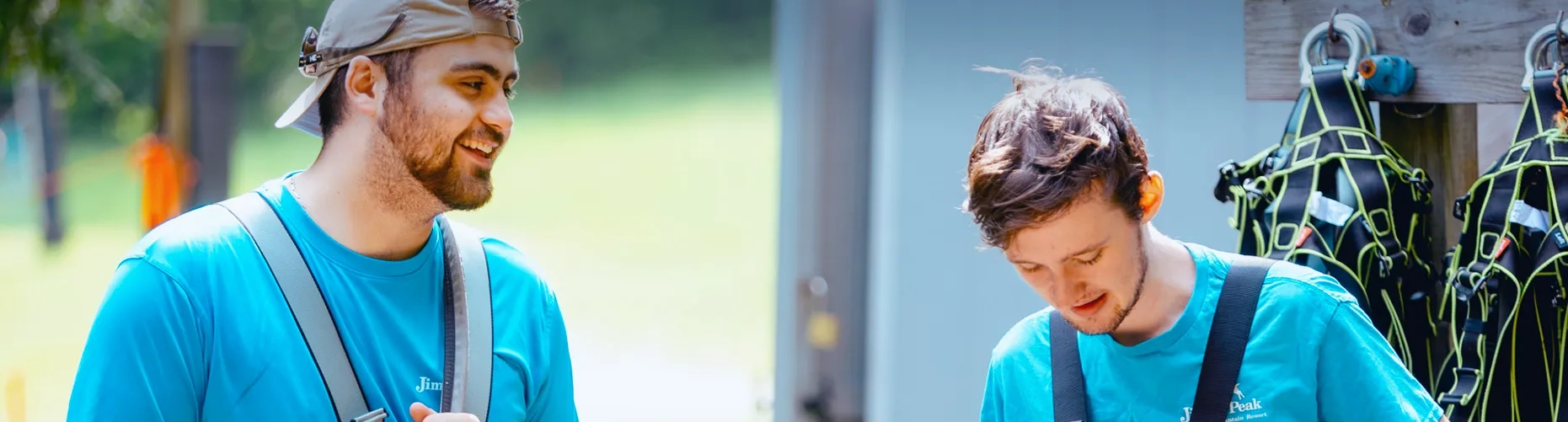 Two Jiminy Peak summer employees in teal staff shirts smiling near a wall of harness equipment outdoors