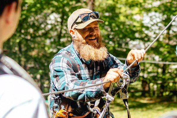 Smiling Jiminy Peak zipline instructor with beard and cap adjusting a cable harness outdoors in summer
